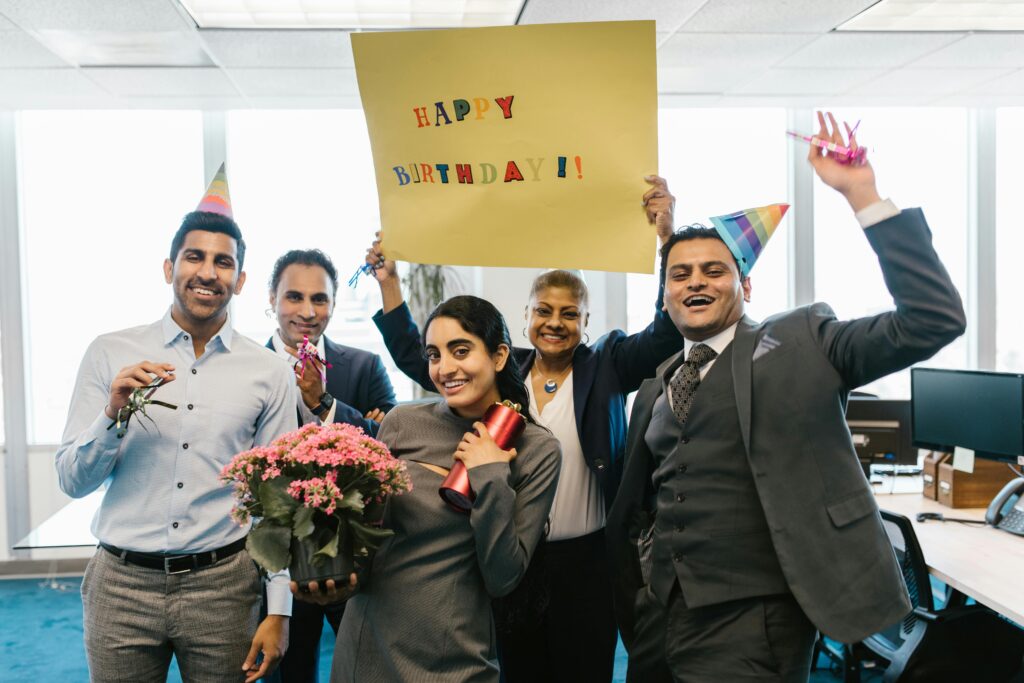 Happy colleagues celebrating a birthday in an office with hats and balloons.